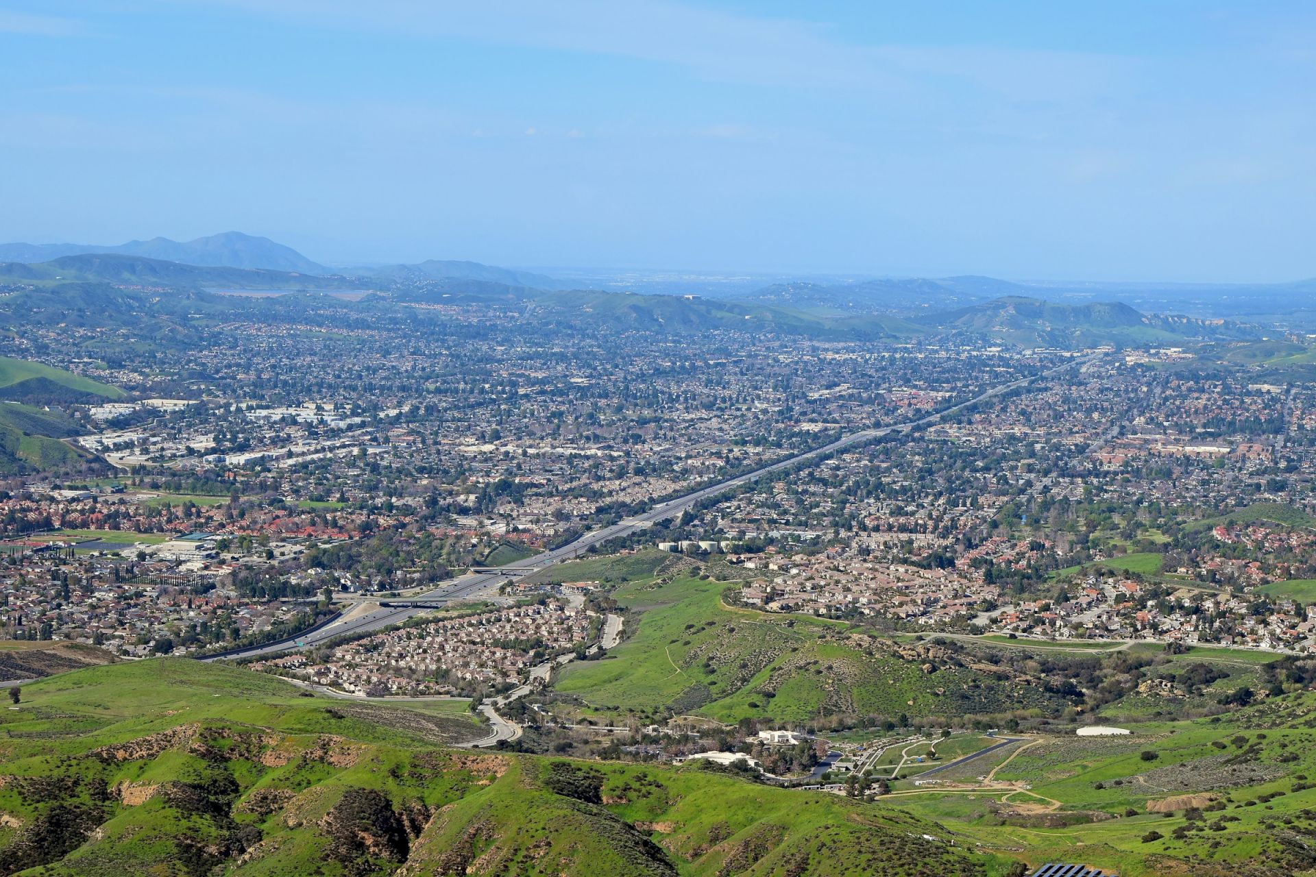 An aerial view of a green valley town with winding roads, a long highway, and surrounding mountains under a clear sky.