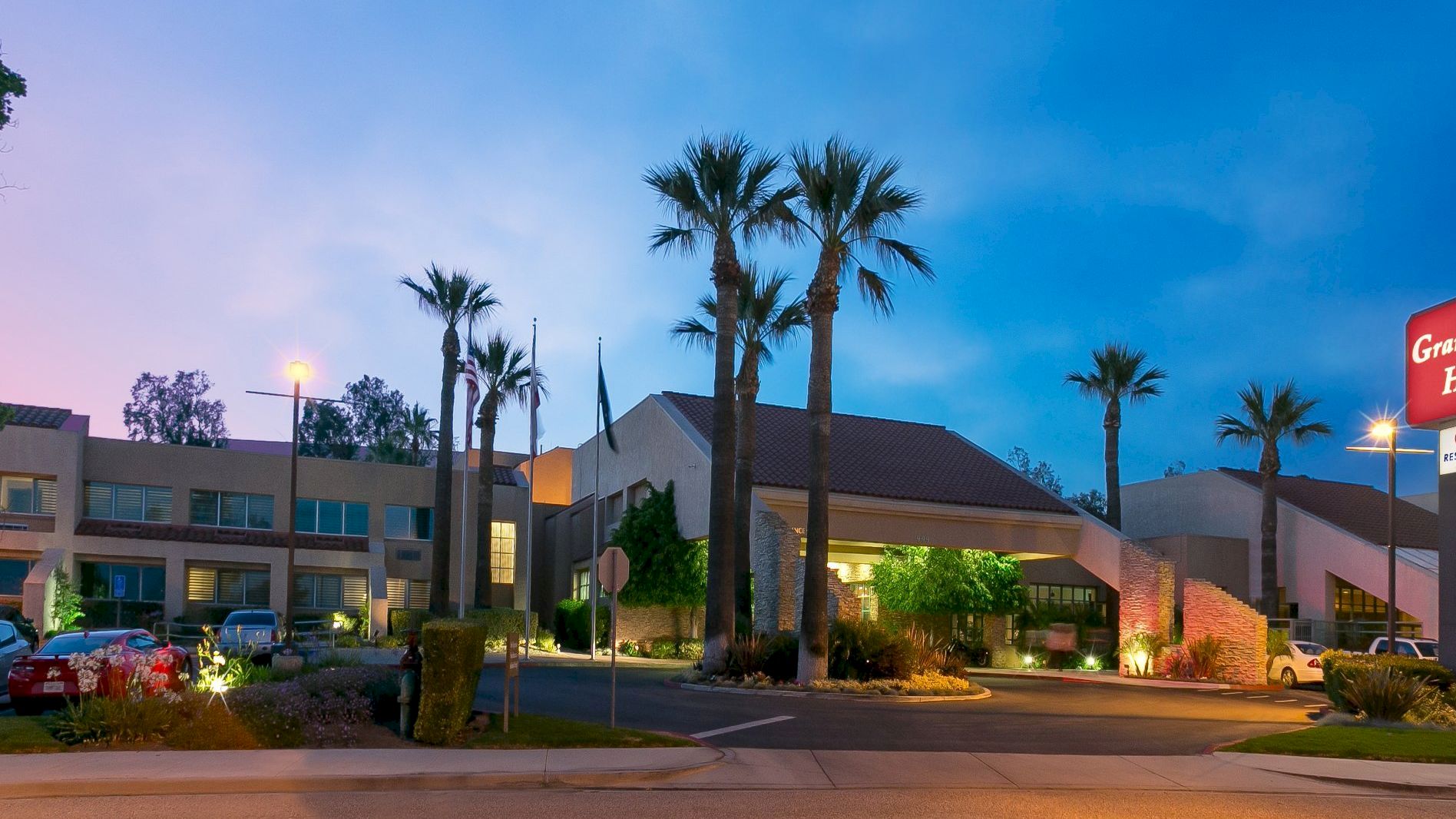 Exterior of the Grand Vista Hotel at dusk, with palm trees, parked cars, and a lit &ldquo;Vistas Restaurant&rdquo; sign.