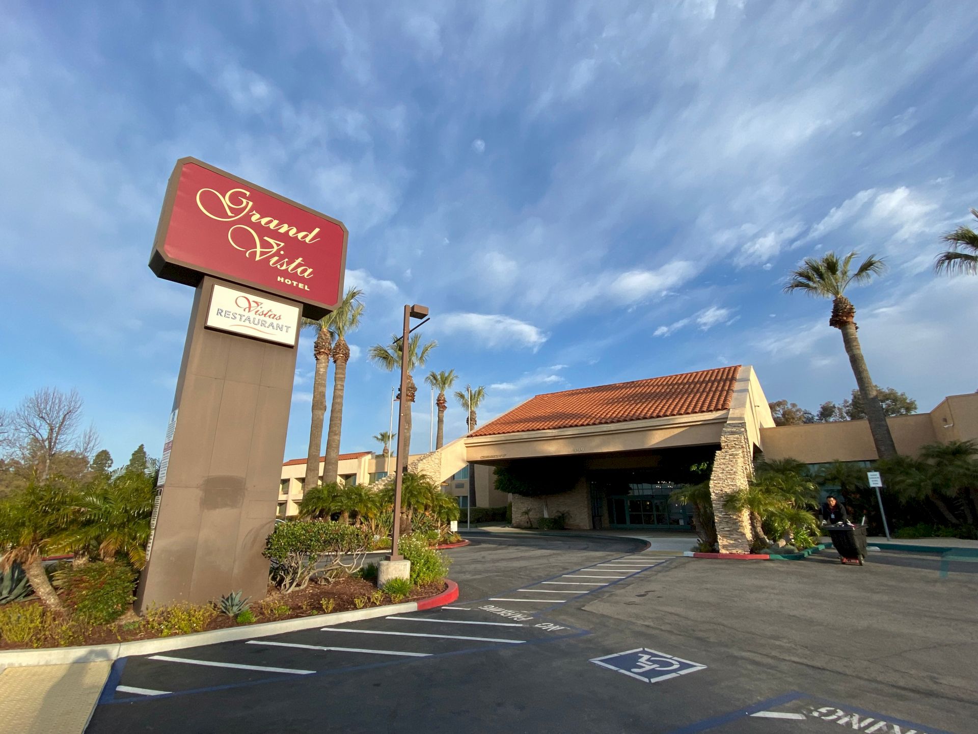 A hotel entrance with a sign reading Grand Vista Hotel and Vistas Restaurant under palm trees and a blue sky.