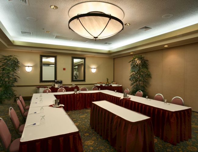 Meeting room with U-shaped tables, red skirting, white napkins, and a central circular chandelier. Plants flank mirrors on beige walls. ending with a period.