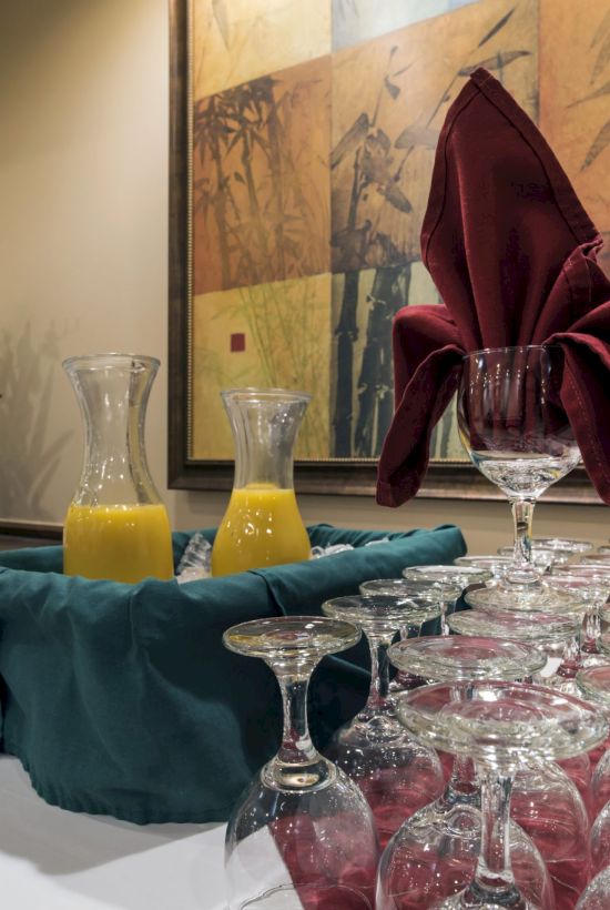 A formal dining setup with a long row of upside-down wine glasses, two carafes of orange juice, folded napkins, and a large framed wall art.