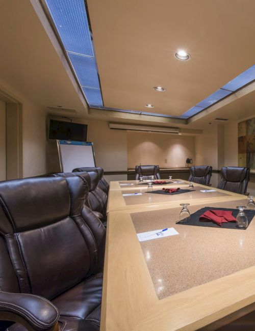 A modern conference room with a large wooden table, black leather chairs, papers and folders scattered, and abstract wall art with soft ceiling lighting.