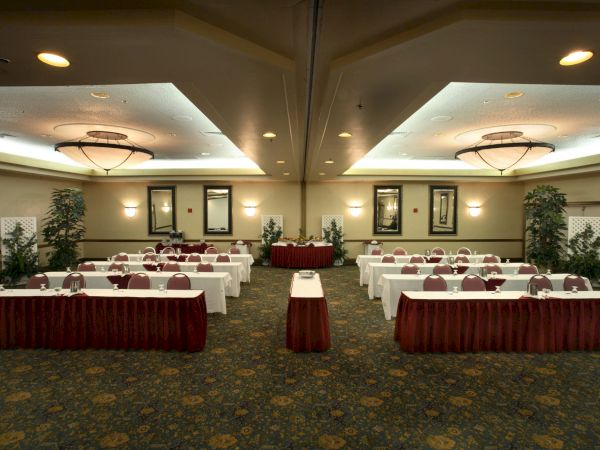 A conference room set up for a meeting with rows of tables and chairs, red table skirts, a central podium, and decorative plants in a well-lit ballroom.