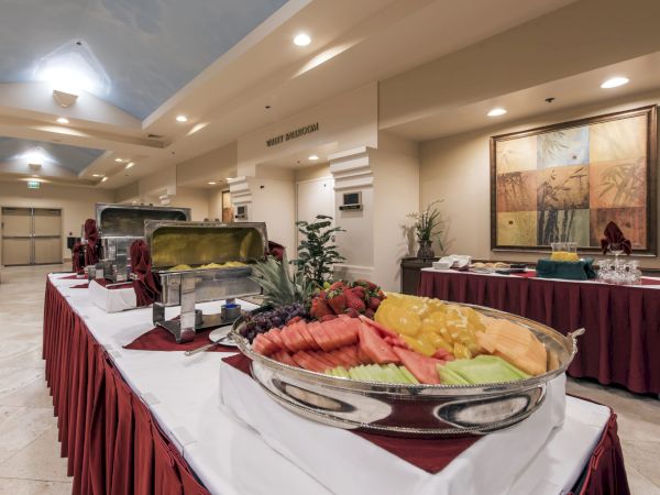 A buffet setup in a hotel banquet hall with chafing dishes, fruit platters, and red-beribboned table skirts, ready for guests.