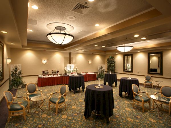A conference or banquet room with round high-top tables, padded chairs, buffet stations, warm lighting, and decorative plants.