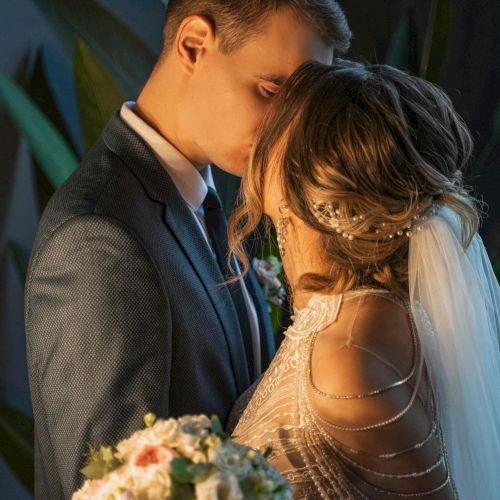 A couple in wedding attire share a tender kiss, the groom in a suit and the bride in a beaded gown, holding a floral bouquet.