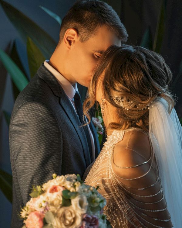 A couple in wedding attire share a tender kiss, the groom in a suit and the bride in a beaded gown, holding a floral bouquet.