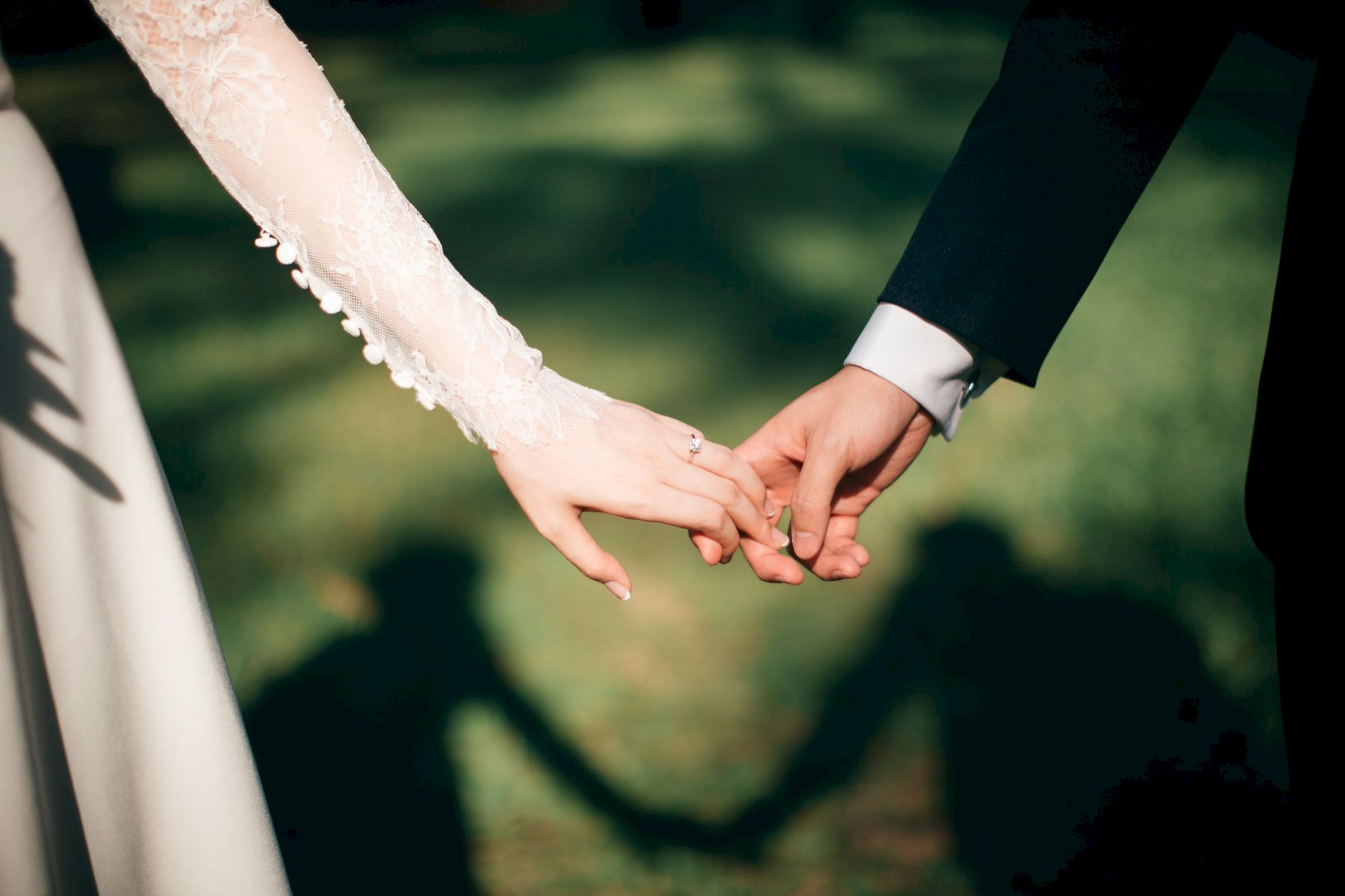 Two people hold hands, wedding attire visible, forming a heart-shaped shadow on a grassy background.