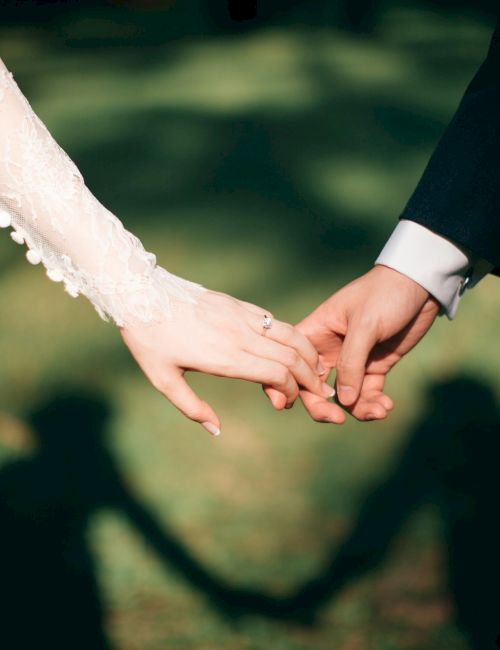 Two hands hold each other, their fingers intertwined, symbolizing a wedding or vows, with a green, blurred outdoor background.