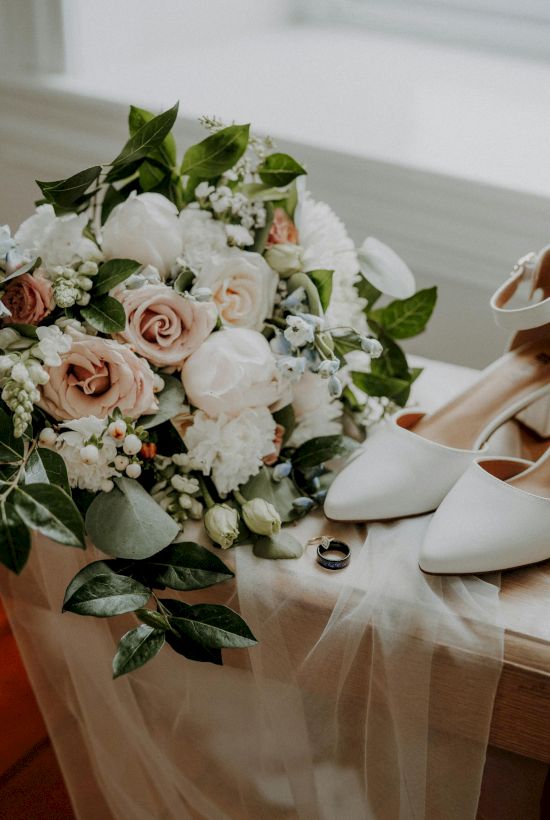 A bridal bouquet of blush pink and white roses with greenery sits beside white heels on a sheer table runner.