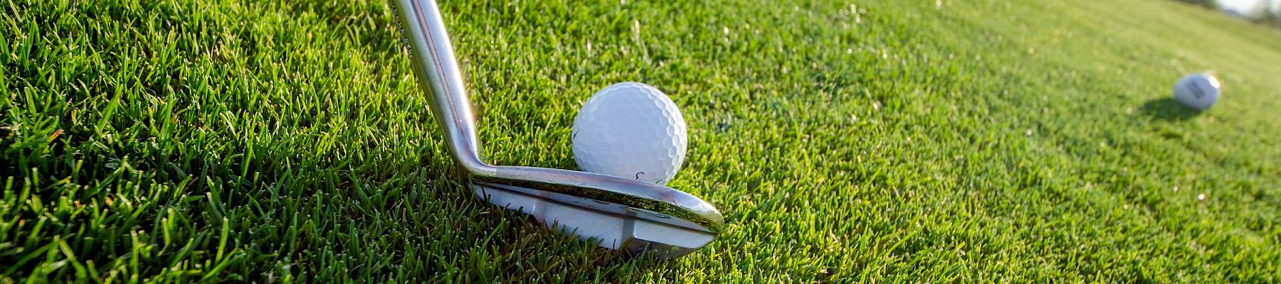 A golf ball sits on a tee about to be struck by a club on a grassy course, with another ball and distant scenery in the background.