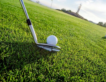 A golf ball sits on a tee about to be struck by a club on a grassy course, with another ball and distant scenery in the background.