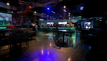 A dimly lit bar or nightclub with scattered tables, neon blue/purple lighting, and a central open floor; screens and a stage area glow in the background.
