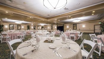 A banquet hall set for a formal event with round tables, white linens, folded napkins, glassware, and white chairs; soft lighting and decor.