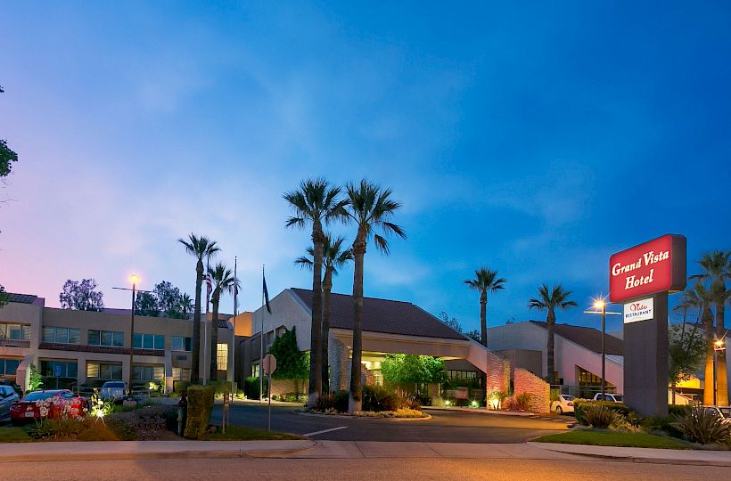 A roadside hotel at dusk with palm trees, a few parked cars, and a lit sign. The sky is blue, and the building glows warmly.