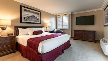 A cozy hotel room with a king bed, burgundy accents, two nightstands, a wall-mounted TV, and a dresser, plus a window seat by the window.