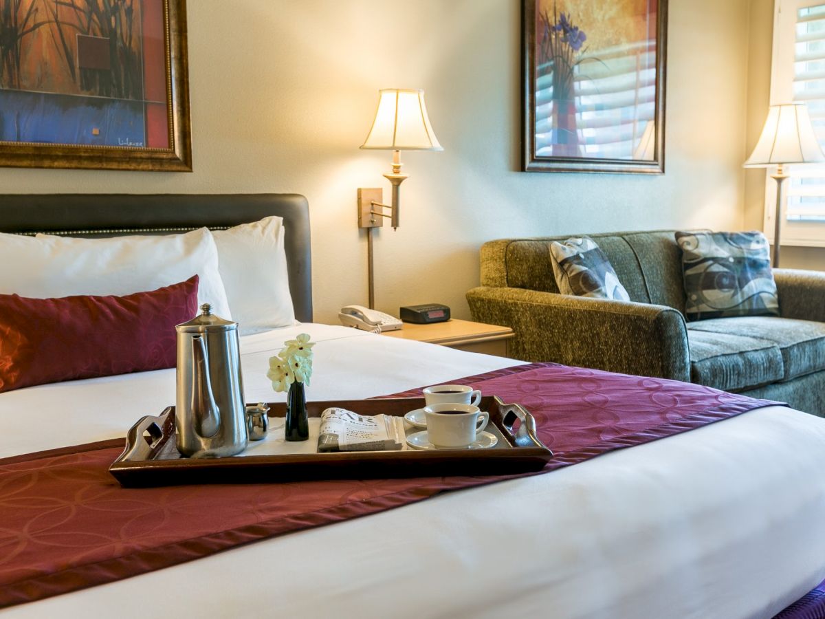 A hotel room with a large bed, burgundy accents, a tray with a kettle and cups, a sofa, lamps, and framed art on the walls.