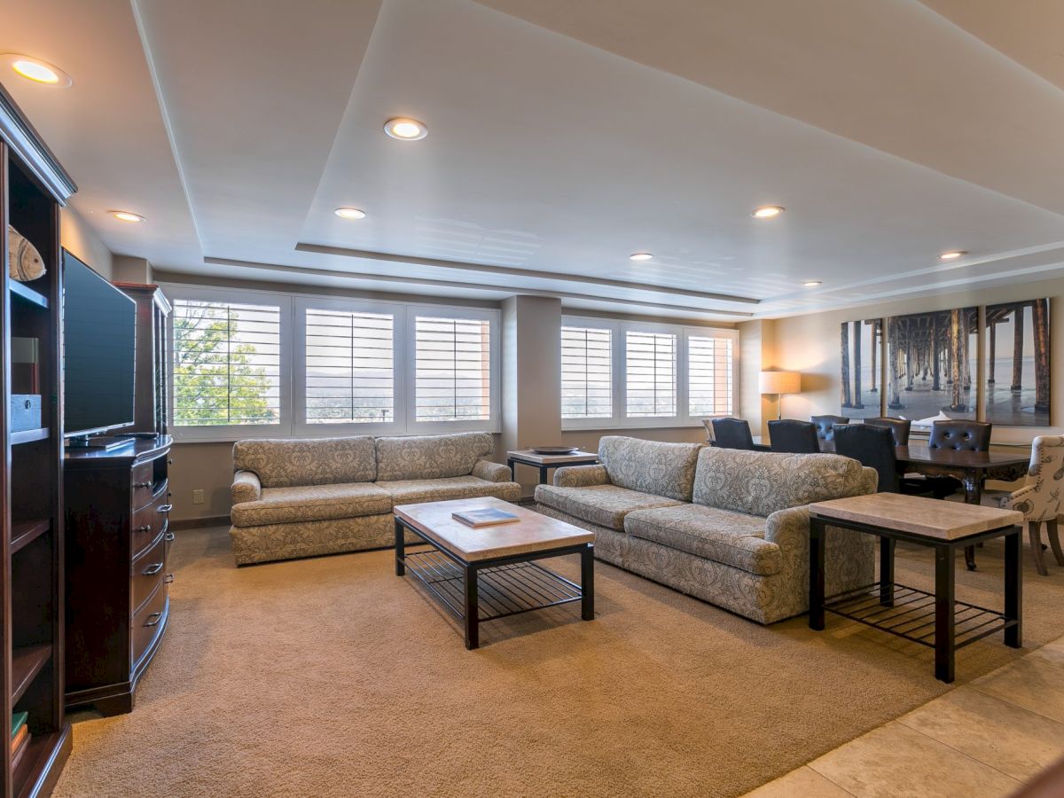 A spacious living room with beige carpet, two matching sofas, a coffee table, side tables, a TV cabinet, and large windows letting in light.