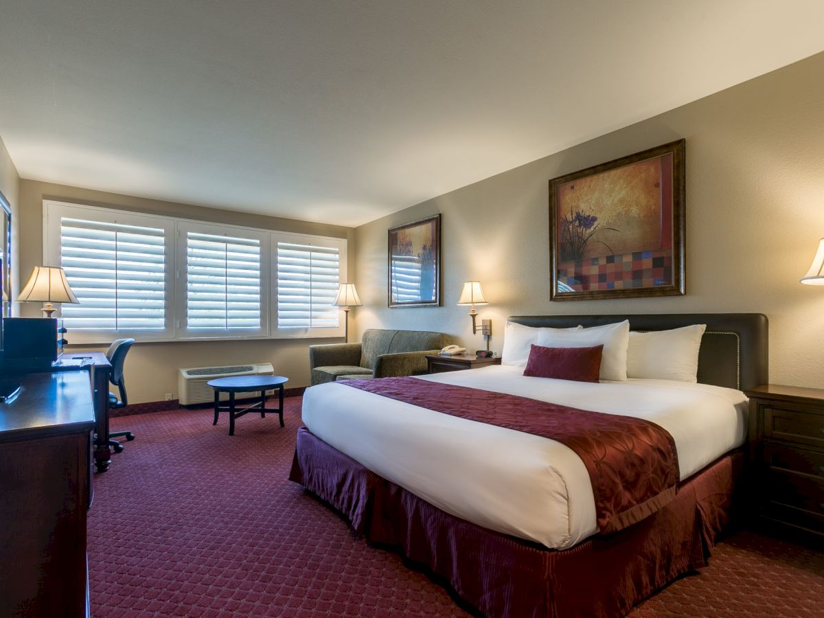 A hotel room with two queen beds, burgundy bed runners, nightstands, a desk, a chair, a TV, and warm lighting in a carpeted suite.
