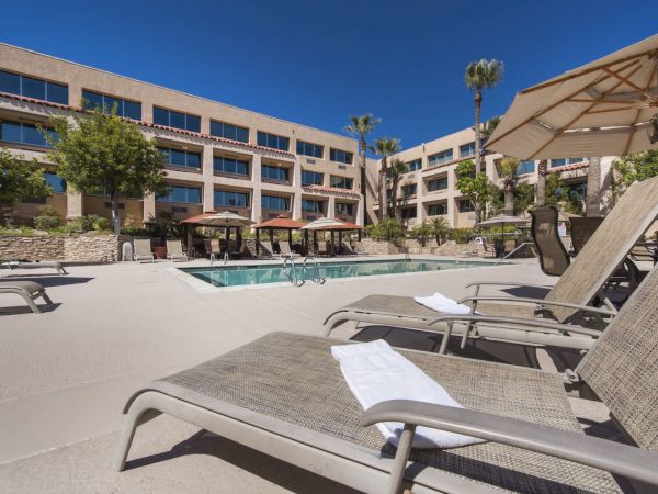 A sunny hotel pool area with lounge chairs, towels, umbrellas, palm trees, and a large beige building in the background.