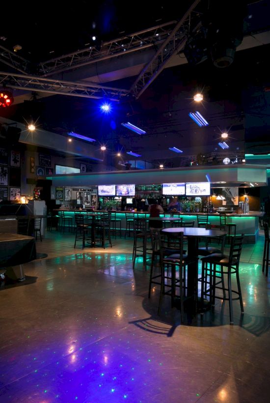 A dimly lit bar or club with neon floor lights, scattered tables and stools, a stage area, and multiple screens and posters around the walls.