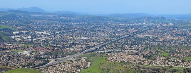 A panoramic hillside town with red-tiled roofs and scattered houses, stretching toward the valley under a clear blue sky.