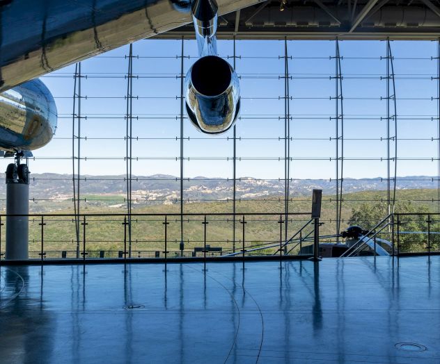 A large aircraft hangar with a jet&rsquo;s tail and open nose gear, view of runway and landscape beyond through a floor-to-ceiling grid window.