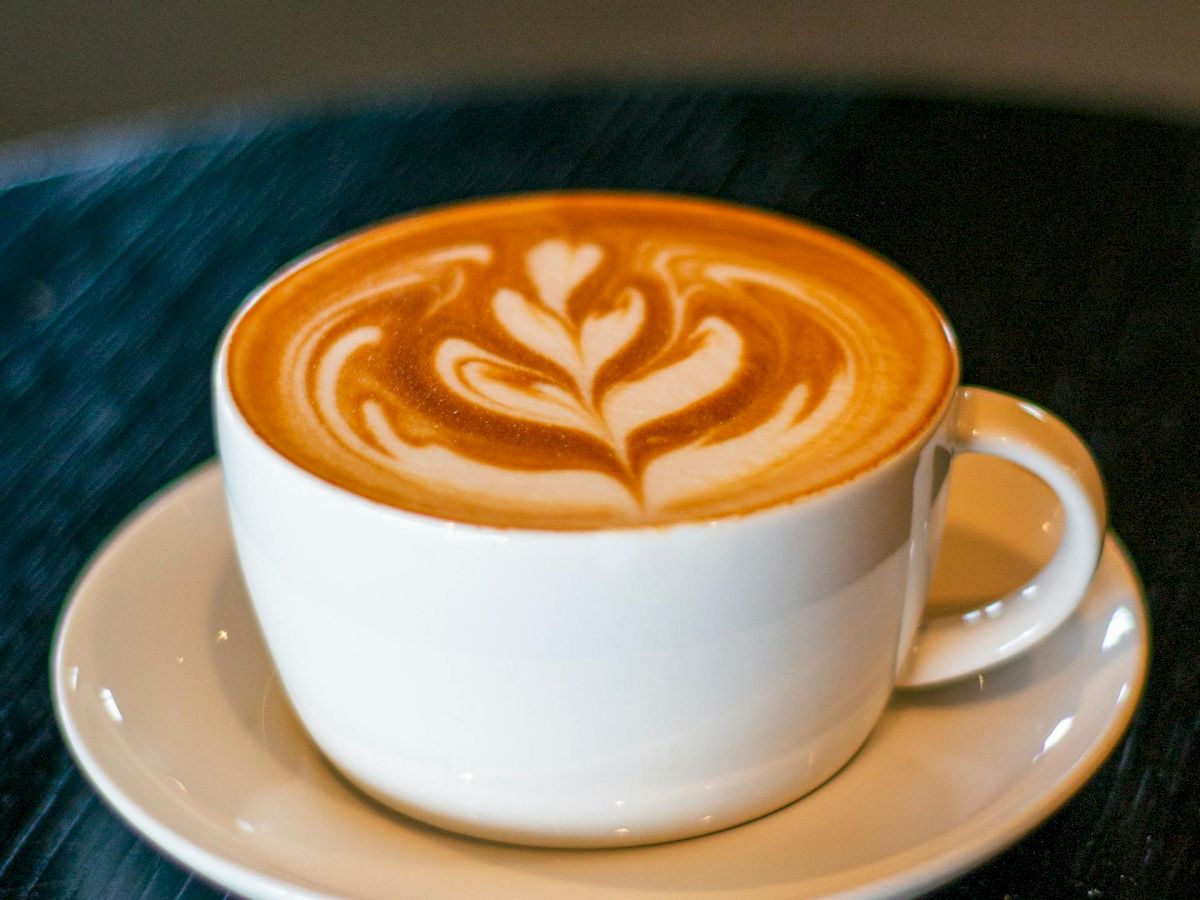 A white cup of latte with latte art on a saucer, resting on a dark table, cozy coffee shop vibe.