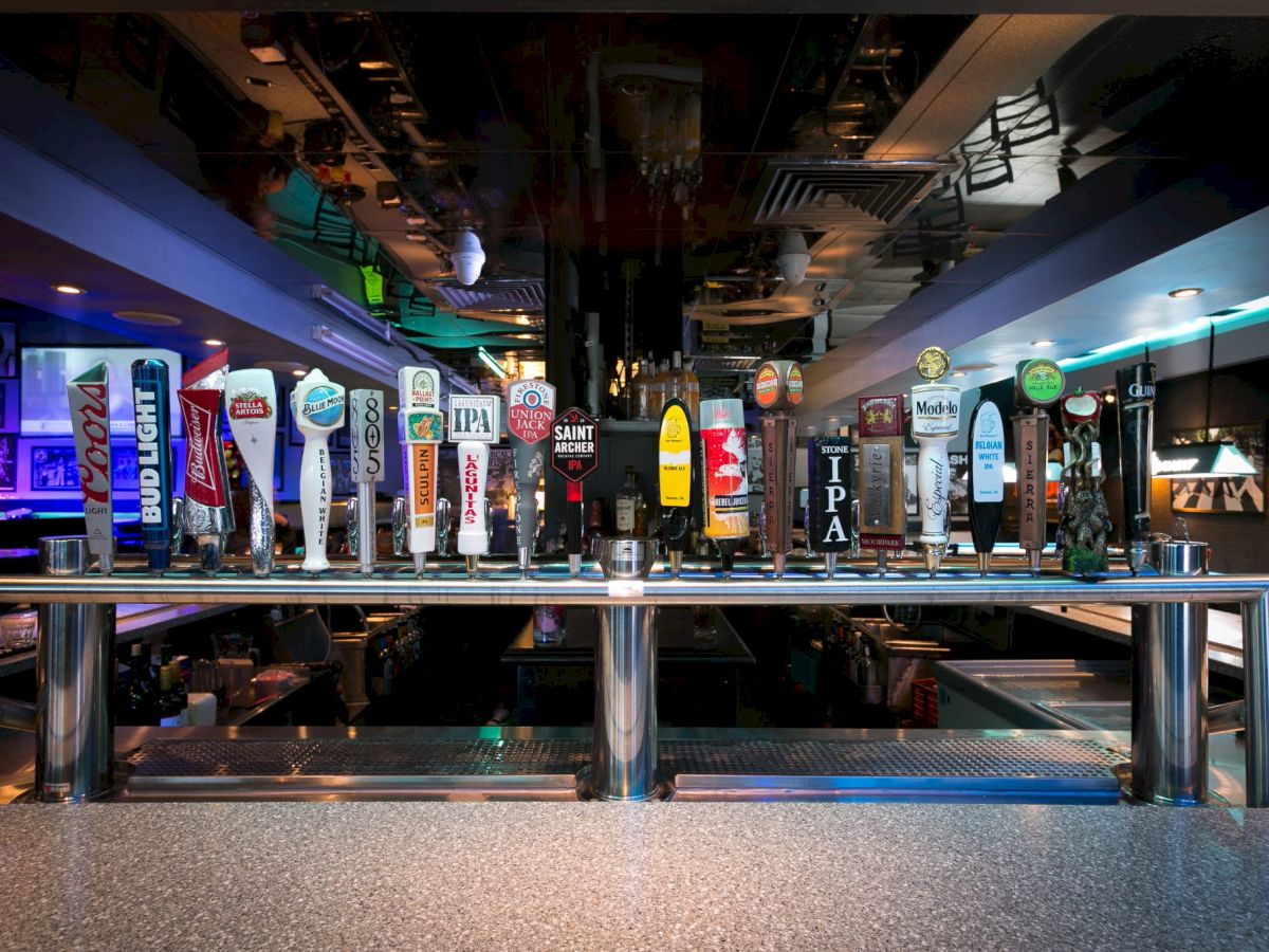 A row of colorful beer taps and signage line a bustling bar or brewery, seen from behind a metal railing.