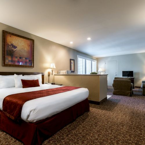 A spacious hotel room with a large bed, burgundy bed runners, bedside lamps, a seating area, desk, and a TV across a patterned carpet, all under soft lighting.