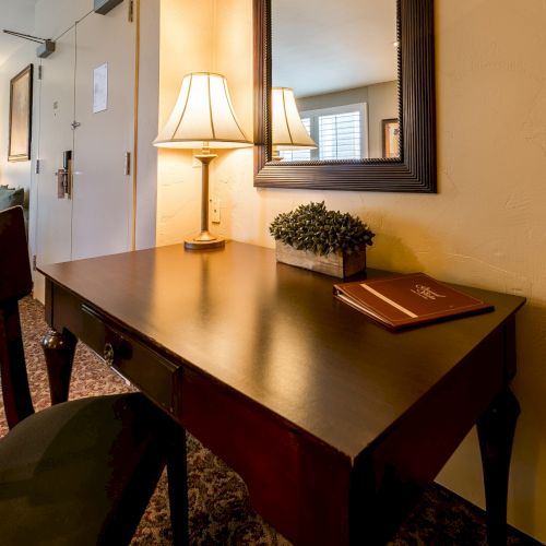 A hotel desk with a lamp, mirror, small plant, and a notebook on a dark wooden table in a cozy room, with a sofa and hallway in view.