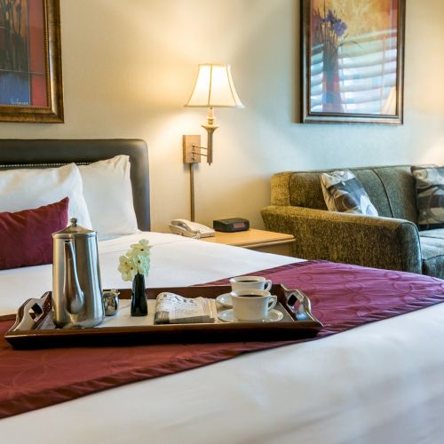 A hotel room with a neatly made bed, a tray of coffee and mugs, and a sitting area with two sofas and lamps, all in warm lighting.