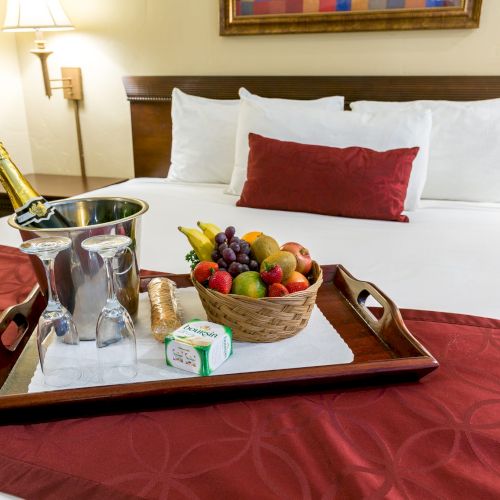 A hotel bed with a tray holding a fruit basket, champagne bucket, glasses, snacks, and a red decorative pillow on a red bed runner.
