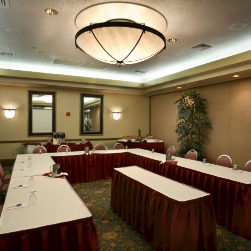 A conference room set up for a meeting with U-shaped tables, burgundy skirting, chairs, and soft lighting.