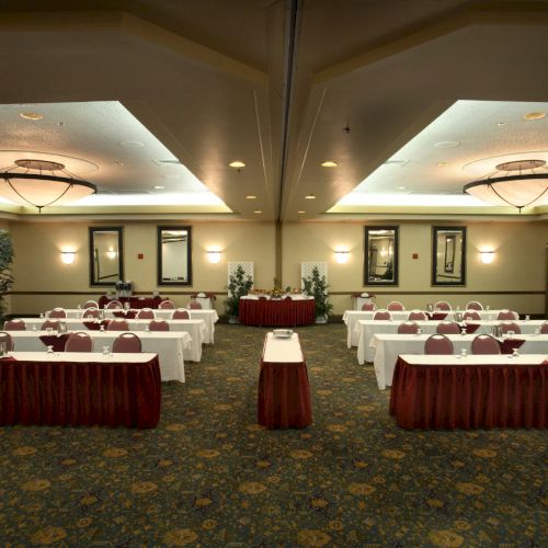 A conference room set for a meeting: long tables with white tablecloths, red skirting, chairs, plants, and a central podium, ready for attendees.
