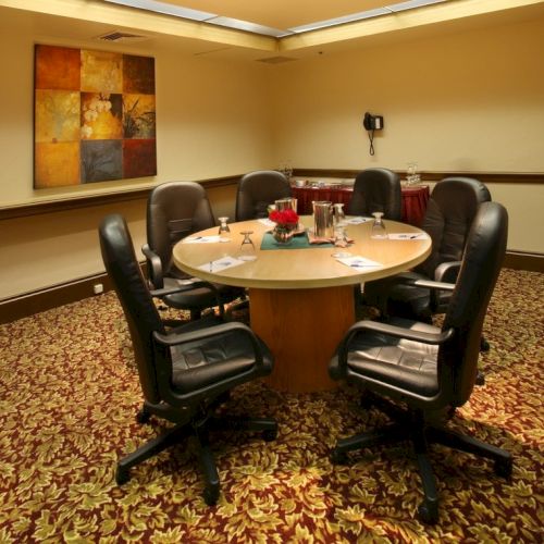 A round conference table with six black office chairs in a carpeted room, a painting on the wall, and a few water bottles and notepads on the table.