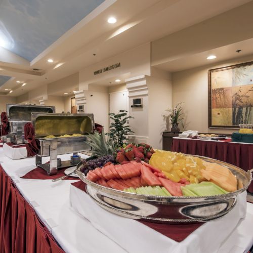 A buffet setup with chafing dishes, fresh fruit platter, and a long table with maroon tablecloths in a banquet hall.