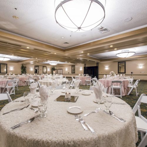 A banquet hall set for a formal event with round tables, white linens, folded napkins, glassware, and white chairs, ready for dining.