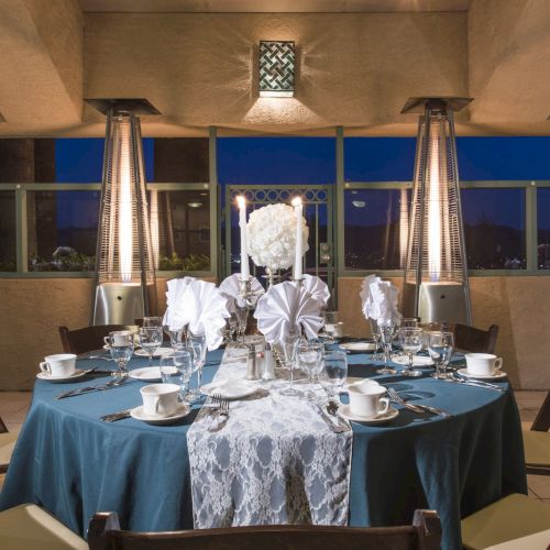 A round dining table set for a formal event with a blue tablecloth, white floral centerpiece, neatly arranged place settings, and four heater lamps on a balcony at night.