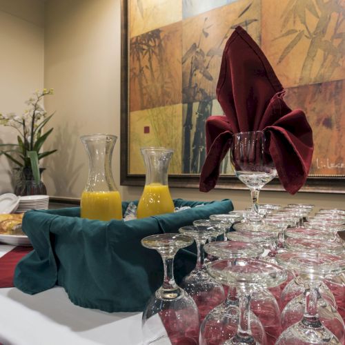 A formal dining setup with a row of upside-down glasses, a red napkin folded like a bow, orange juice carafes, and a framed background.