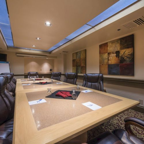 A modern conference room with a large oval table, black leather chairs, papers and folders, and abstract wall art in a well-lit, professional setting.