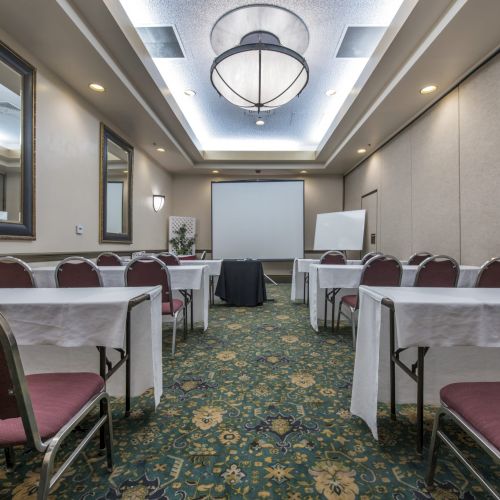 A conference room set for a presentation with long tables, red chairs, a projector screen, flip chart, and a patterned carpet.