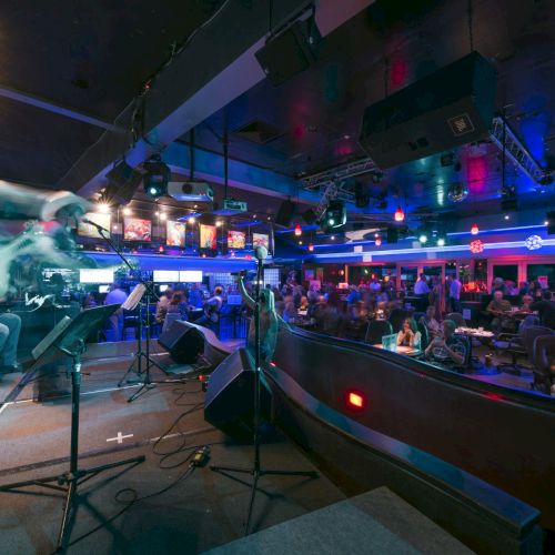 A live band rocks a small venue with guitars and amps on stage, an intimate audience, neon lights, and a cozy bar area nearby.