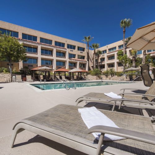 A modern hotel pool area with lounge chairs, umbrellas, and a rectangular pool, surrounded by a tan building and palm trees.