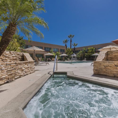 A resort-style pool area with a hot tub in the foreground, stone walls, palm trees, and sun loungers under umbrellas.