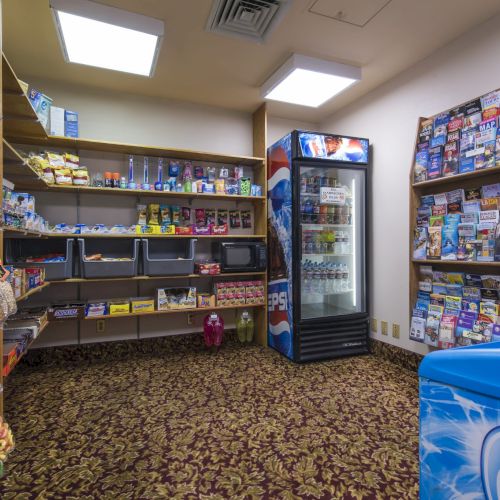 A small convenience shop: shelves with snacks, a drink fridge, brochures on racks, a blue cooler in the foreground, and a patterned carpet.