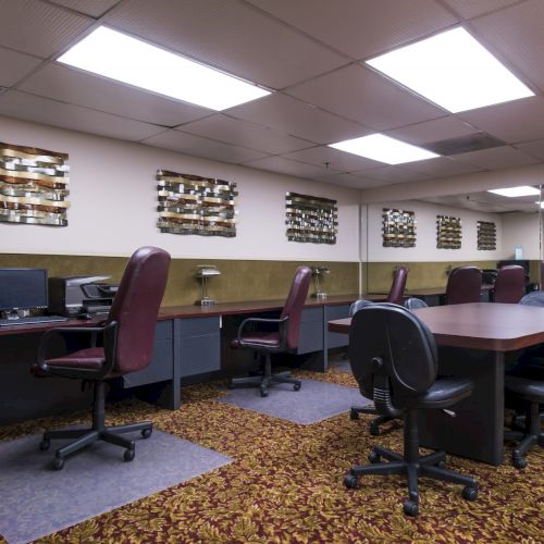 A dim office lab with several desks and computers, maroon chairs, and a long table in a carpeted room with wood-paneled walls and fluorescent lights.