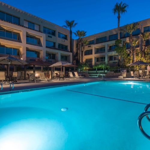 A bright hotel pool at dusk with blue water, lounge chairs, palm trees, and a multi-story tan building surrounding the area. Top it at 140 characters, always ending the sentence.
