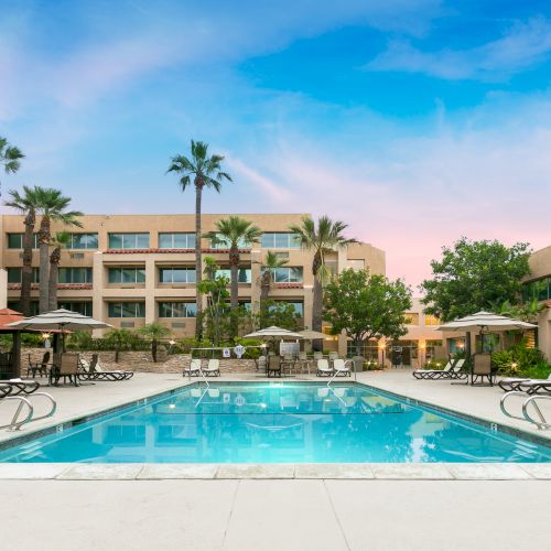 A sunny hotel pool area with lounge chairs, palm trees, and a three-story beige building surrounding the pool, under a blue sky.