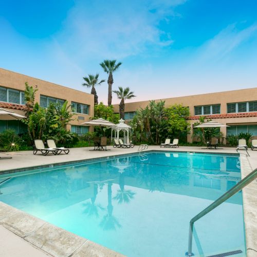 A hotel-style courtyard with a sunny pool, lounge chairs, umbrellas, palm trees, and a two-story beige building surrounding the courtyard.